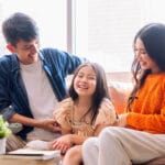 A smiling mother, father, and young daughter sit together on a couch in a bright living room, enjoying each others company—peace of mind made possible with ประกันสุขภาพเหมาจ่าย. A small green plant and books are on the table in front of them.