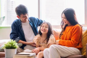 A smiling mother, father, and young daughter sit together on a couch in a bright living room, enjoying each others company—peace of mind made possible with ประกันสุขภาพเหมาจ่าย. A small green plant and books are on the table in front of them.